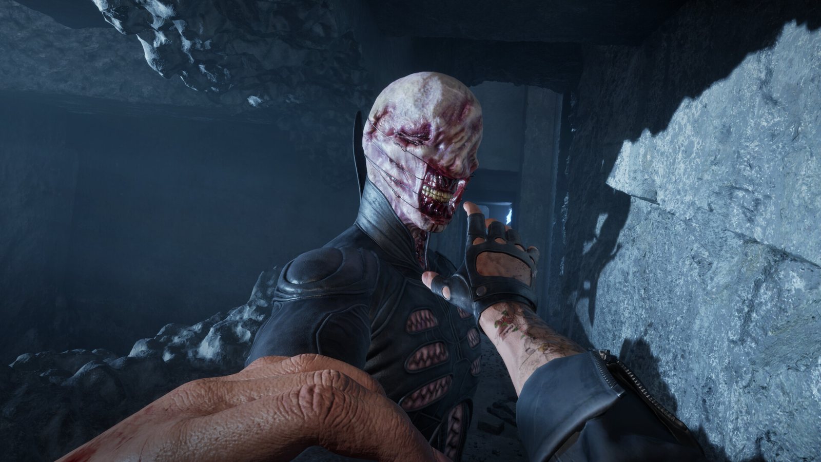 A first-person hand reaches toward a pale stitched figure with a torn, tooth-filled mouth in a rocky cave. Official screenshot. A first-person hand reaches toward a pale stitched figure with a torn tooth-filled mouth